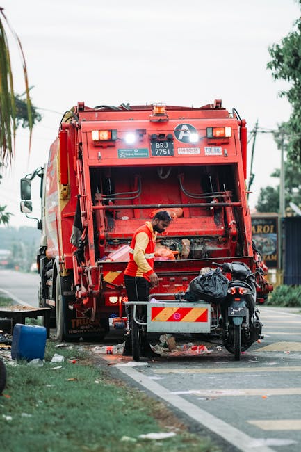 A waste collection truck with a white body and rusted metal components is positioned on a cobblestone street, collecting rubbish from a blue wheeled bin held by a worker dressed in blue overalls and a