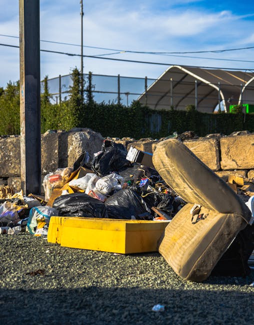 A pile of mixed waste consisting of black plastic rubbish bags, loose paper, cardboard, and assorted debris is situated on a gravel surface beside a stone wall made of large, rough-hewn concrete block