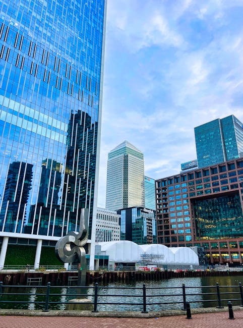The image depicts a modern urban landscape with a body of water in the foreground reflecting the surrounding structures. A red train is seen crossing a dark grey elevated railway bridge that extends a