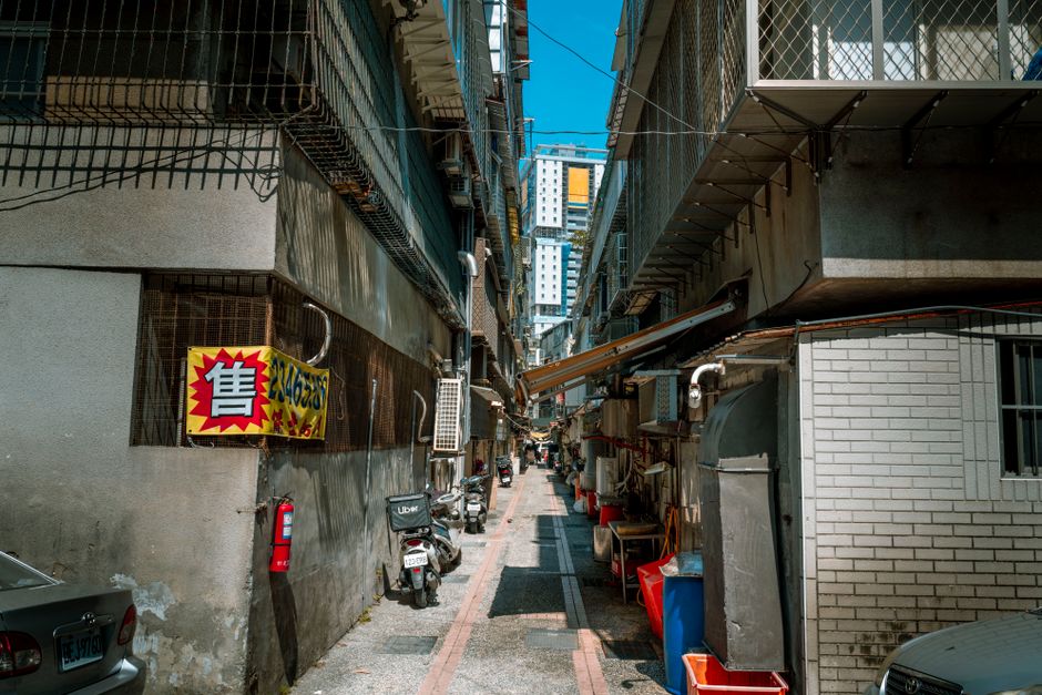 A large, weathered beige rubbish sack made of coarse fabric rests on the pavement beside a brick and concrete wall in a narrow urban alleyway. The sack appears full and slightly sagging, with some pla